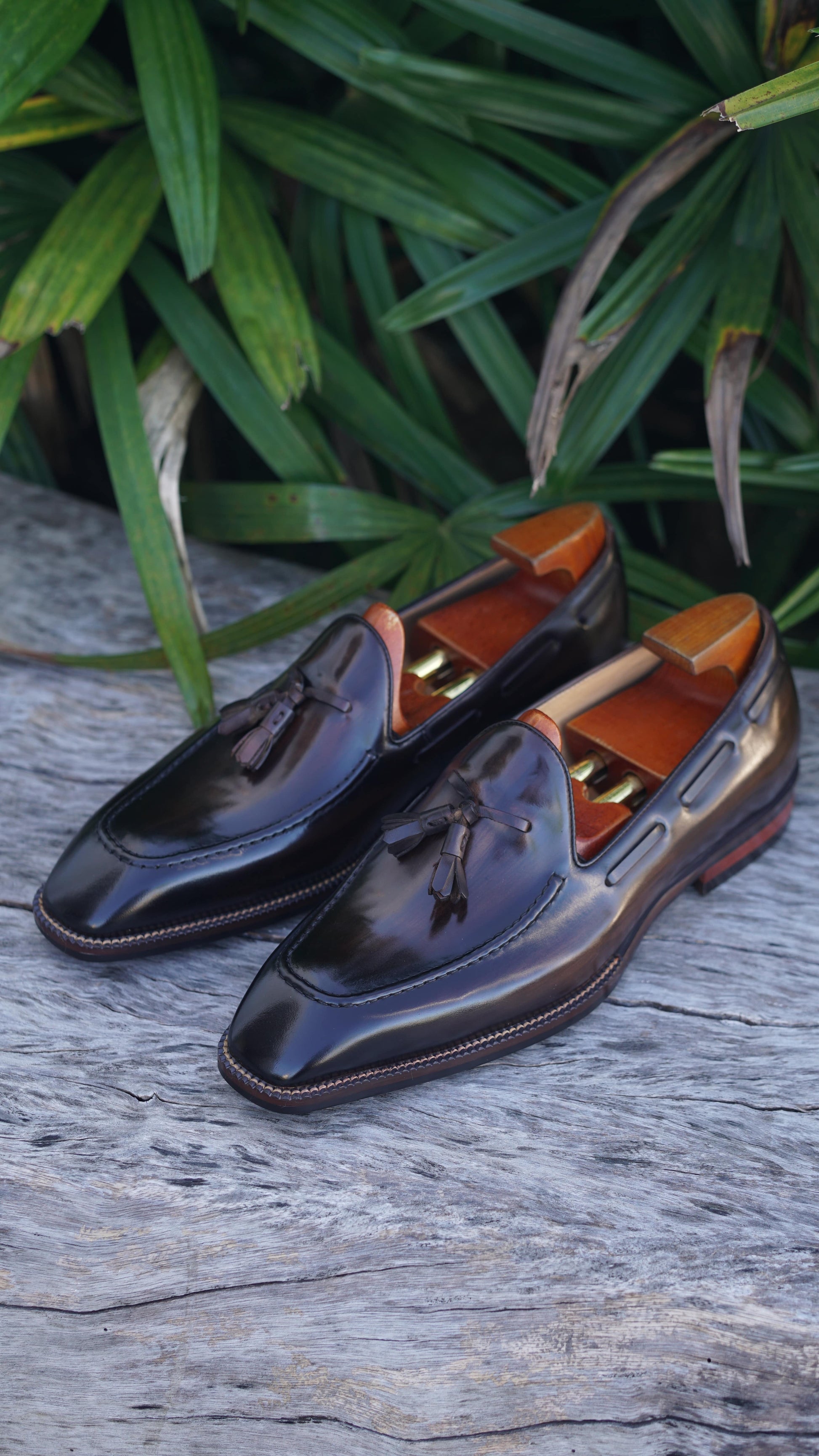 Hand-Made Dark Brown Leather Tassel Loafers – pair with shoe trees on a wooden surface, with green foliage, highlighting sophisticated design.