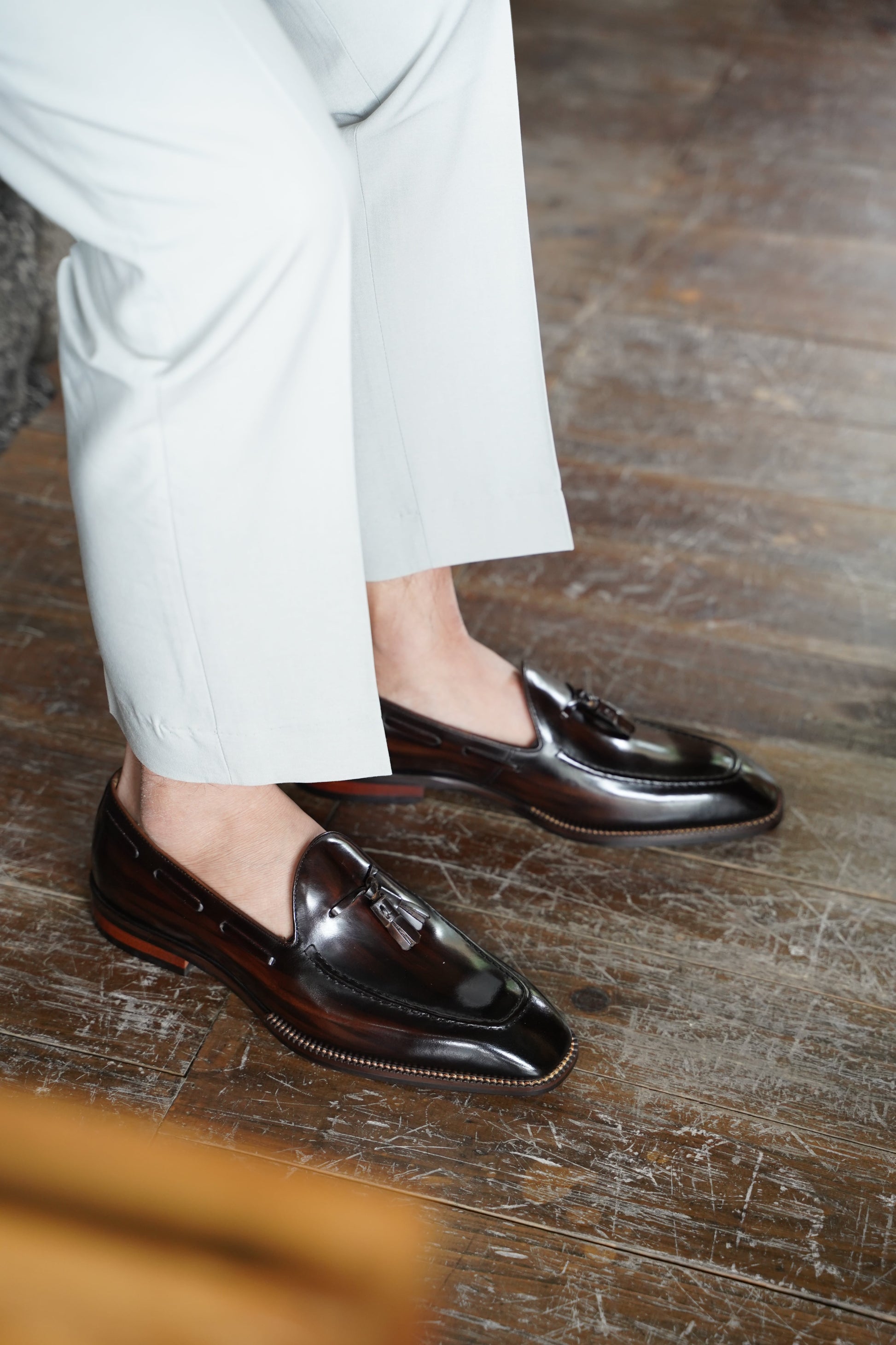 Hand-Made Dark Brown Leather Tassel Loafers – worn by a man in light trousers, showcasing hand-painted patina and sleek almond-toe silhouette on a wooden floor.