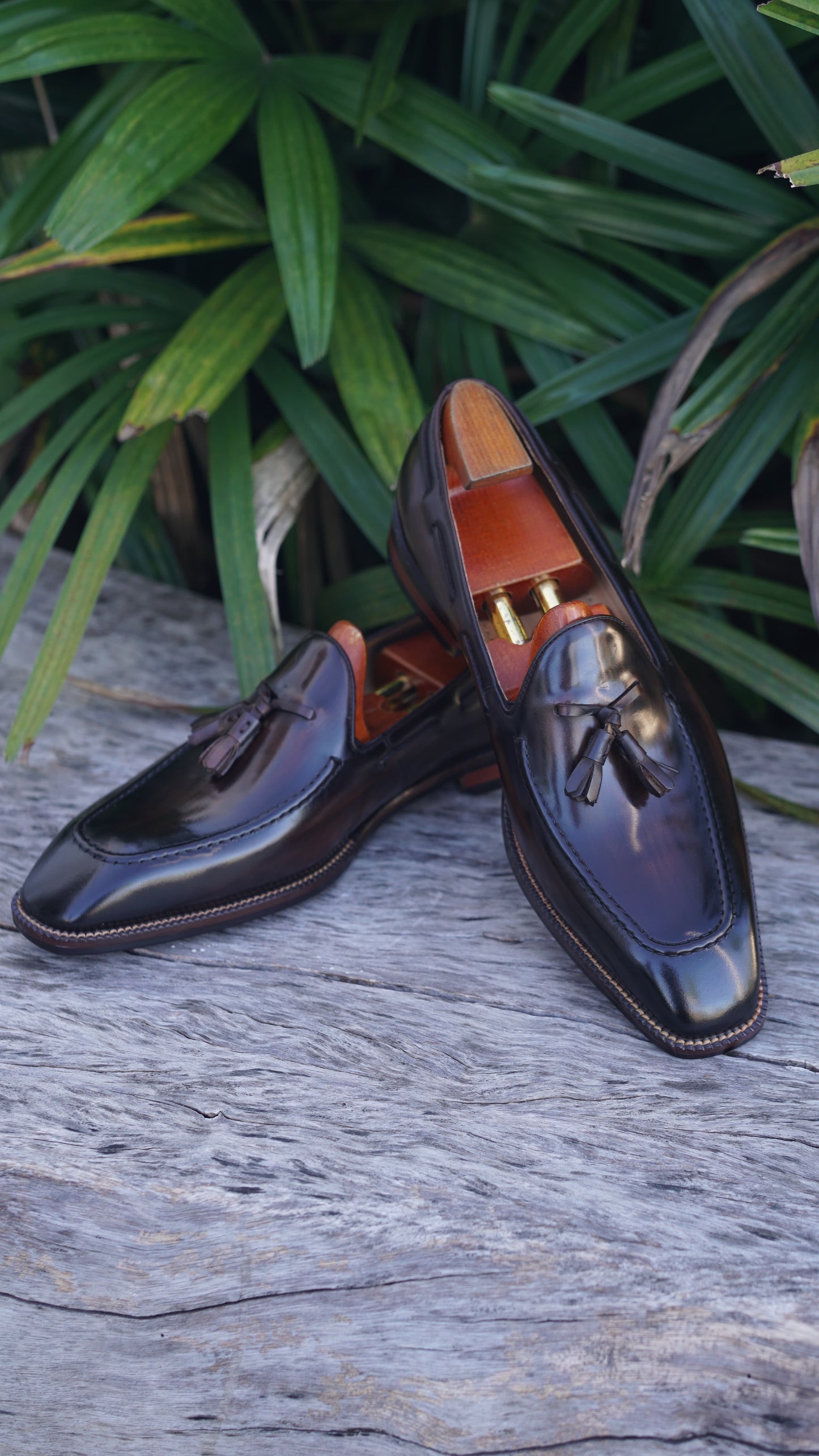 Hand-Made Dark Brown Leather Tassel Loafers – pair with shoe trees, with green foliage in background, highlighting classic tassel detail and hand-painted finish.