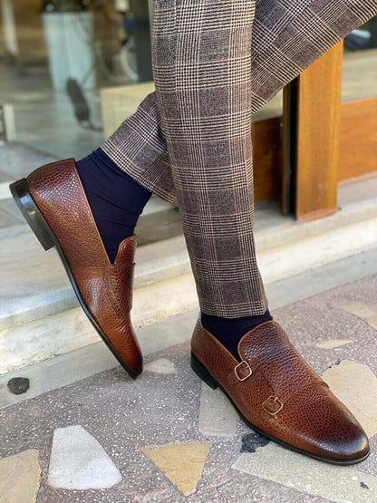 Men's brown grained leather double monk strap loafers styled with plaid trousers and navy socks, walking on a tiled surface.