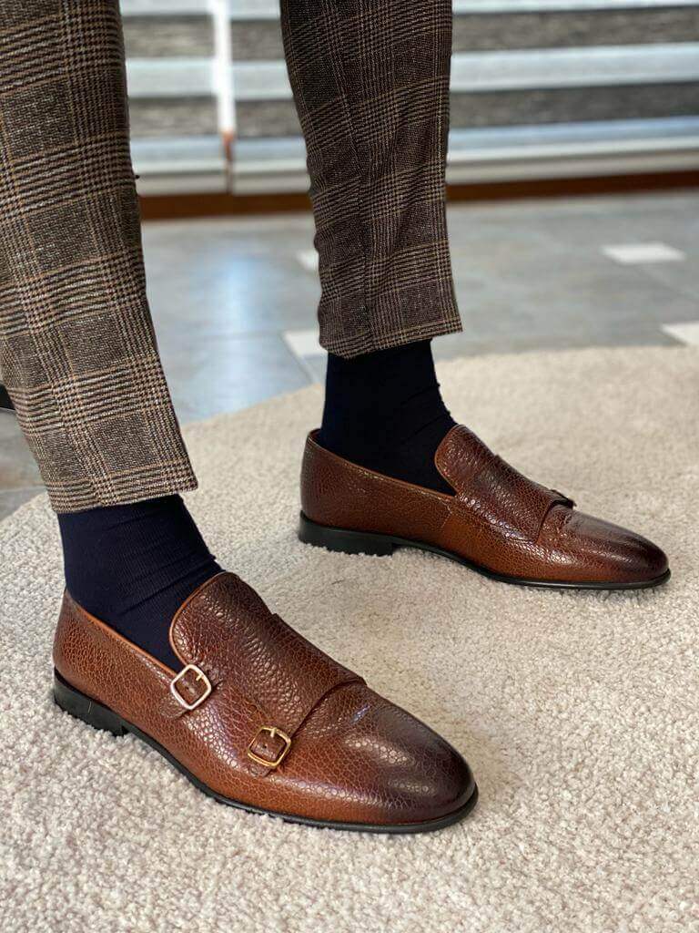 A man's feet in brown grained leather double monk strap loafers and plaid pants, one foot resting on a metal chair leg.
