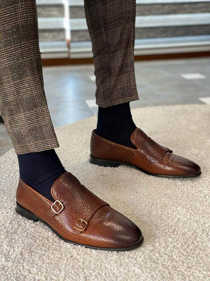 A man's feet in brown grained leather double monk strap loafers and plaid pants, one foot resting on a metal chair leg.