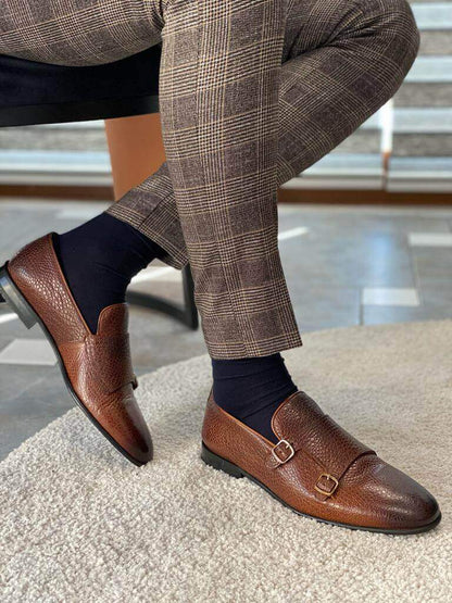 Close-up of men's brown grained leather double monk strap loafers and plaid trousers on a patterned tile and marble floor.