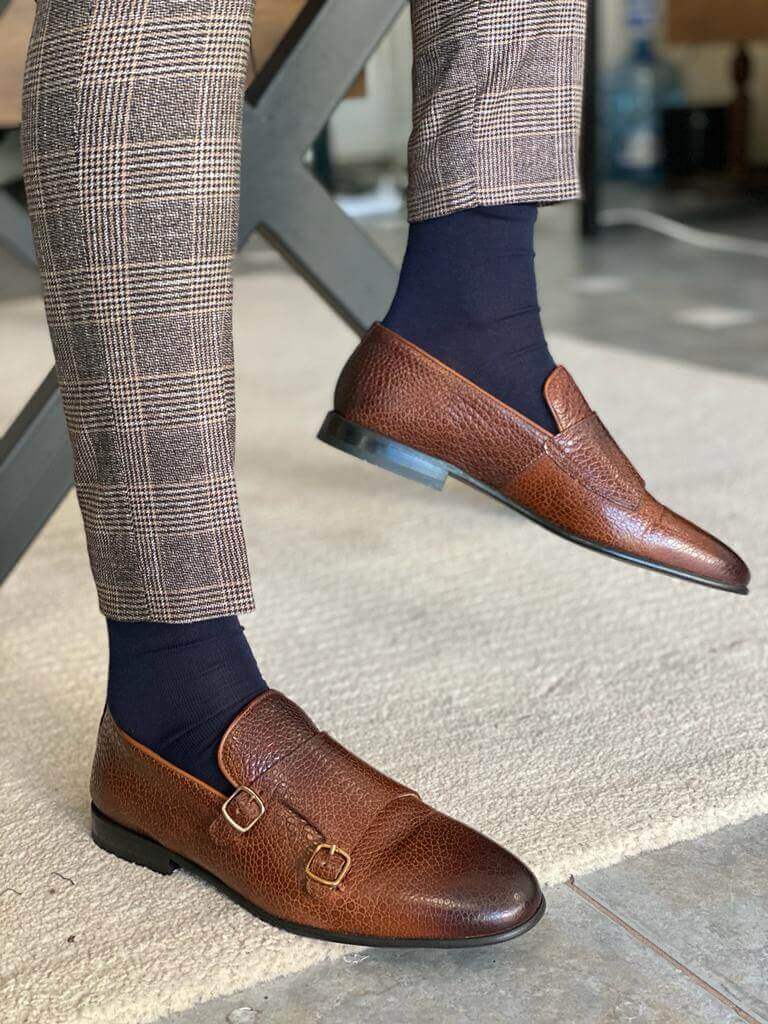 A man wearing brown grained leather double monk strap loafers and plaid trousers, standing on a light patterned rug.