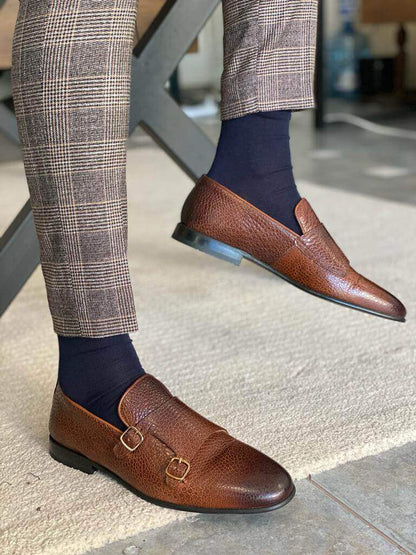 A man wearing brown grained leather double monk strap loafers and plaid trousers, standing on a light patterned rug.