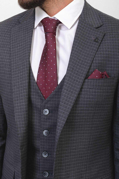 A well-dressed man wearing a Dark Gray Checkered Three-Piece Suit stands confidently in a studio setting. The suit features a subtle checkered pattern, paired with a white dress shirt, a burgundy polka dot tie, and a matching pocket square, adding a touch of elegance. The ensemble is completed with black leather dress shoes, enhancing its classic and refined look. The man has neatly styled hair and a trimmed beard, embodying a polished and sophisticated appearance, ideal for formal and professional occasion