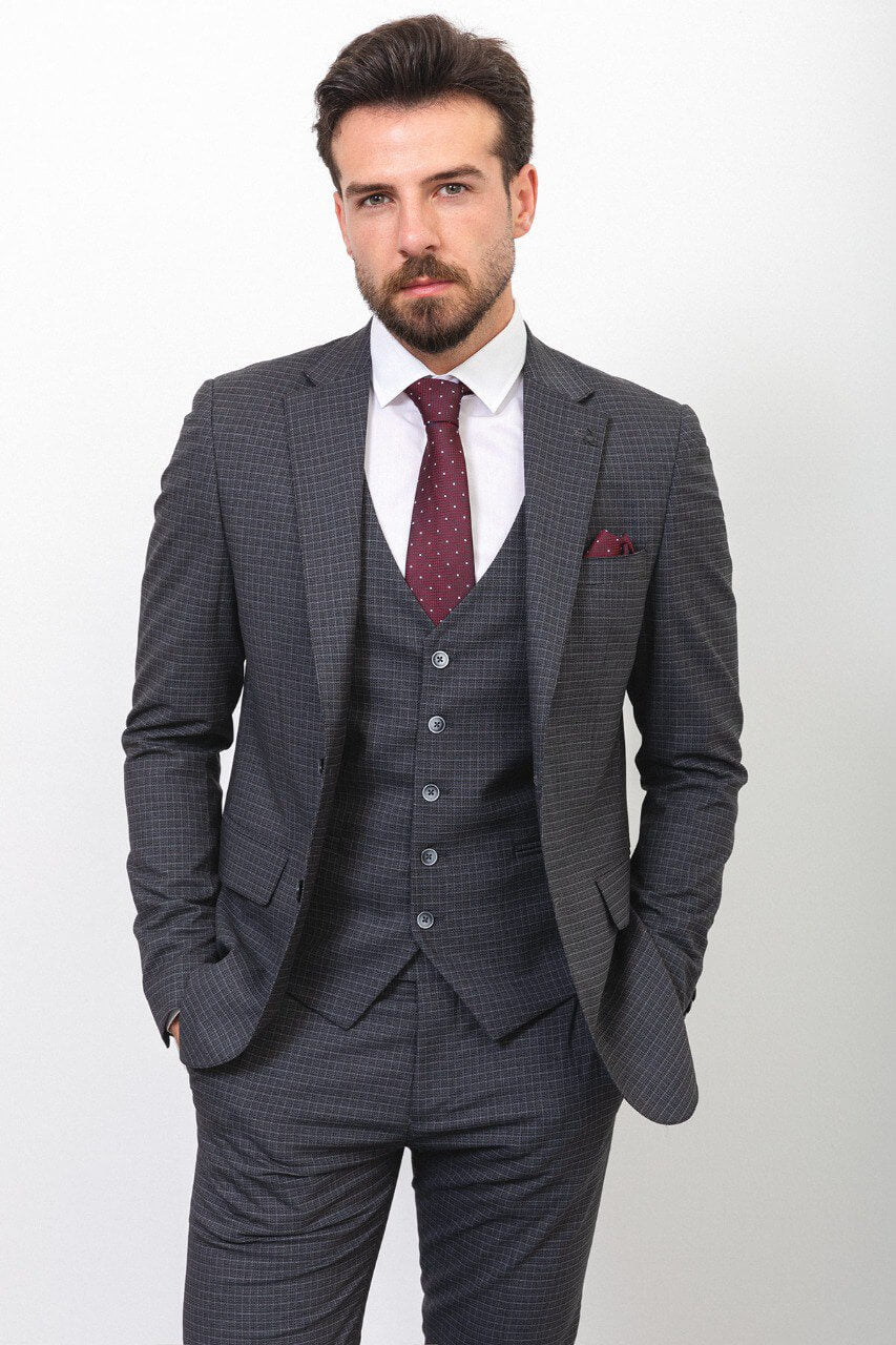 A well-dressed man wearing a Dark Gray Checkered Three-Piece Suit stands confidently in a studio setting. The suit features a subtle checkered pattern, paired with a white dress shirt, a burgundy polka dot tie, and a matching pocket square, adding a touch of elegance. The ensemble is completed with black leather dress shoes, enhancing its classic and refined look. The man has neatly styled hair and a trimmed beard, embodying a polished and sophisticated appearance, ideal for formal and professional occasion