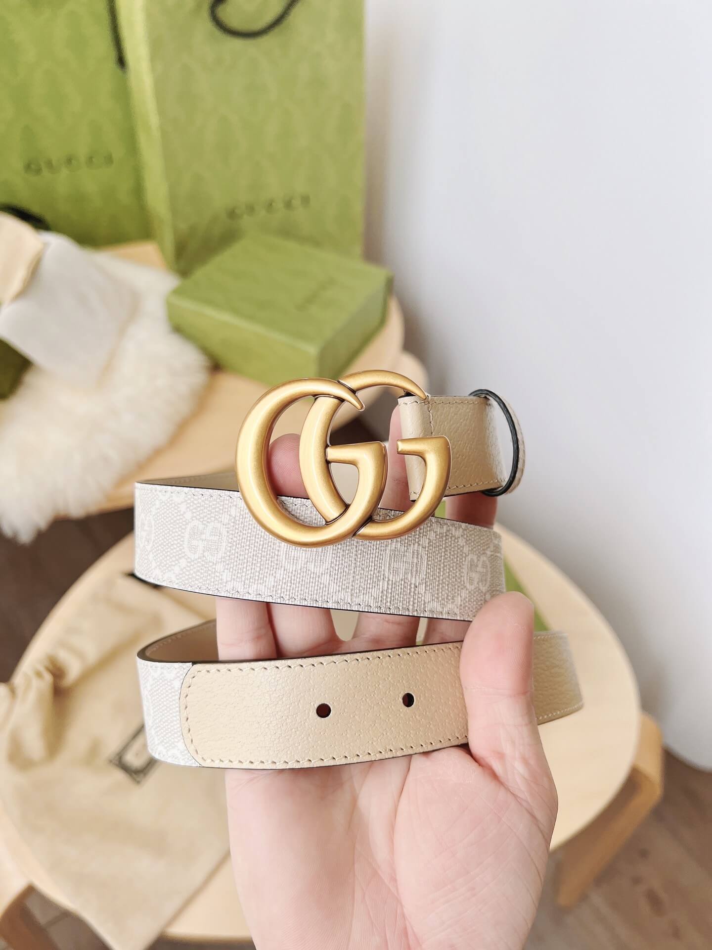 Beige belt with gold Gucci buckle held by a hand, with a green Gucci bag in the background.