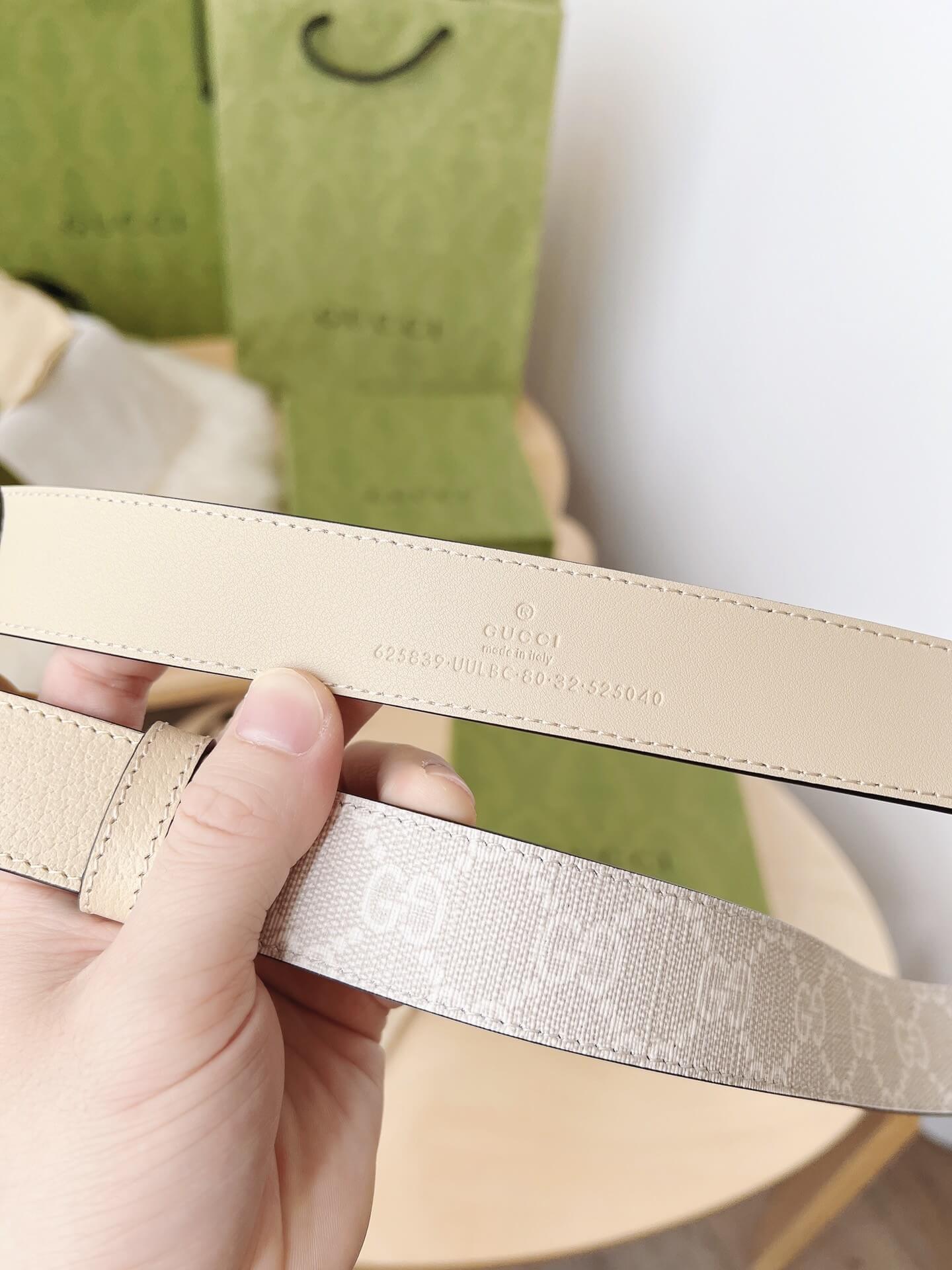 Close-up of a beige Gucci belt held by a hand with a blurred green background.