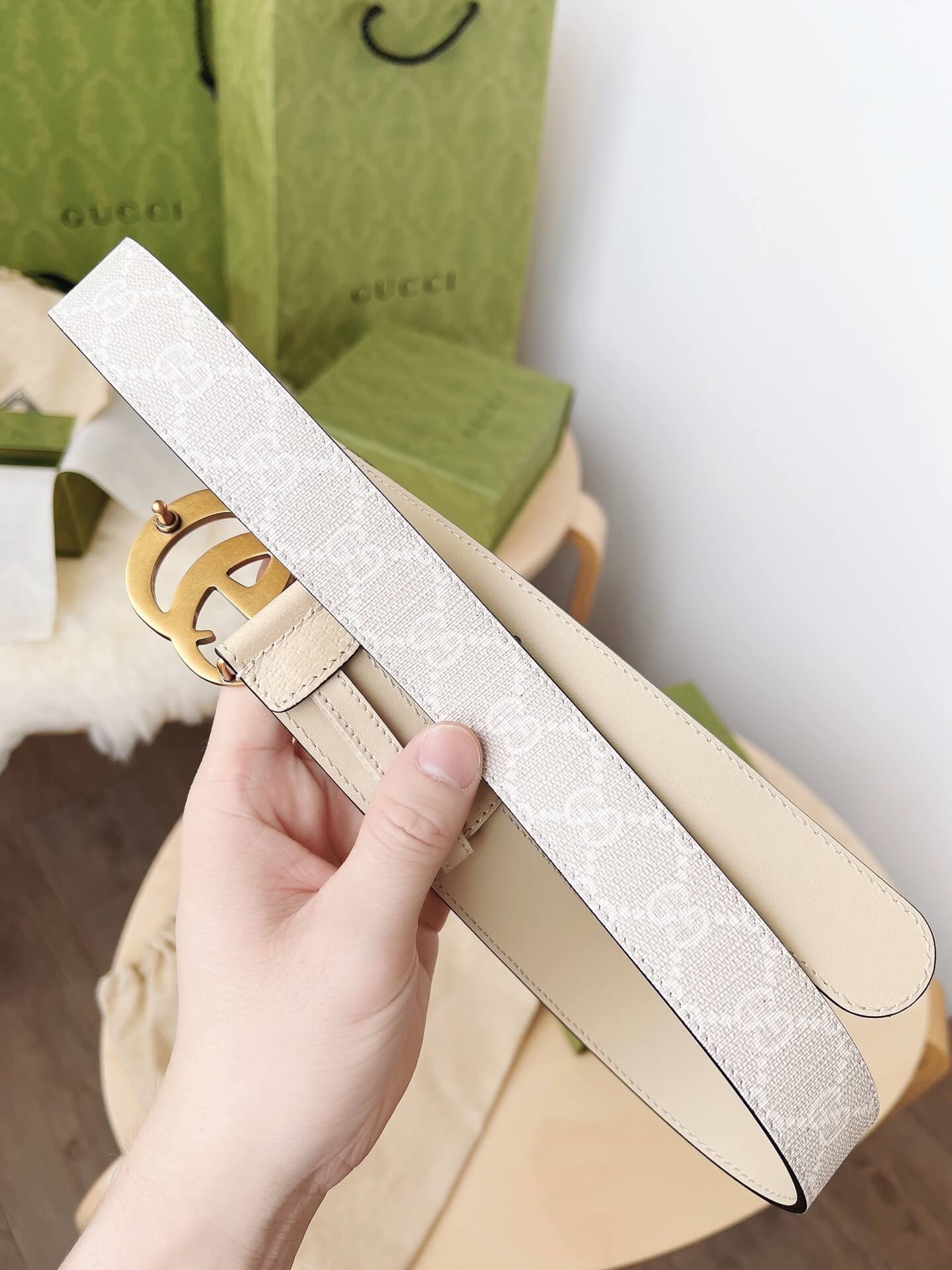 Beige handbag with gold logo handle held by a hand, Gucci boxes in the background