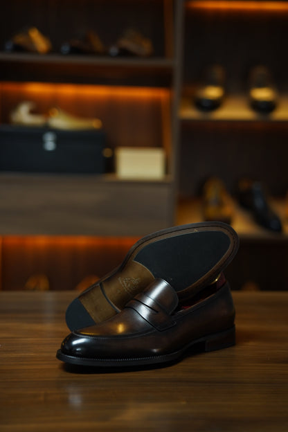 Hand-Burnished Brown Leather Penny Loafers – pair resting on a wooden surface, with one shoe showing the outsole, highlighting the hand-polished finish and comfort.