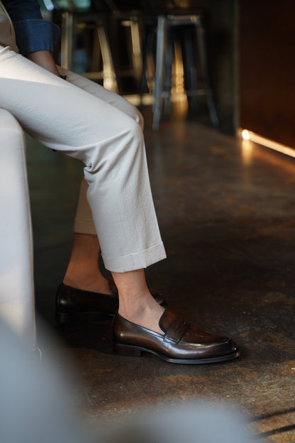 Hand-Burnished Brown Leather Penny Loafers – worn by a man sitting, paired with light trousers and a denim shirt, showcasing seamless comfort and class.
