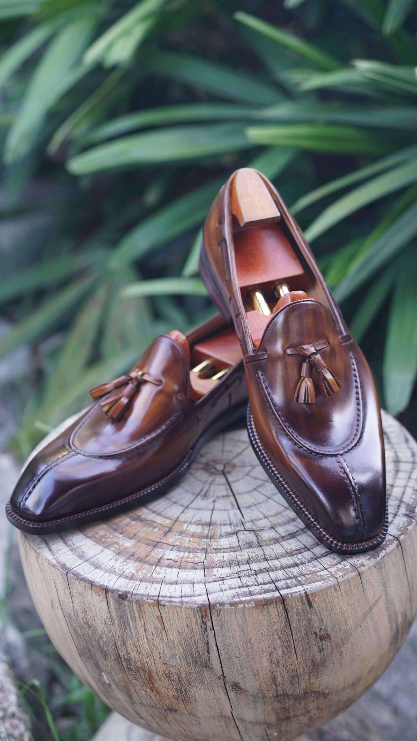 Hand-Burnished Brown Patina Leather Tassel Loafers – pair with shoe trees on a wooden stump, showcasing polished leather and classic tassel detail.