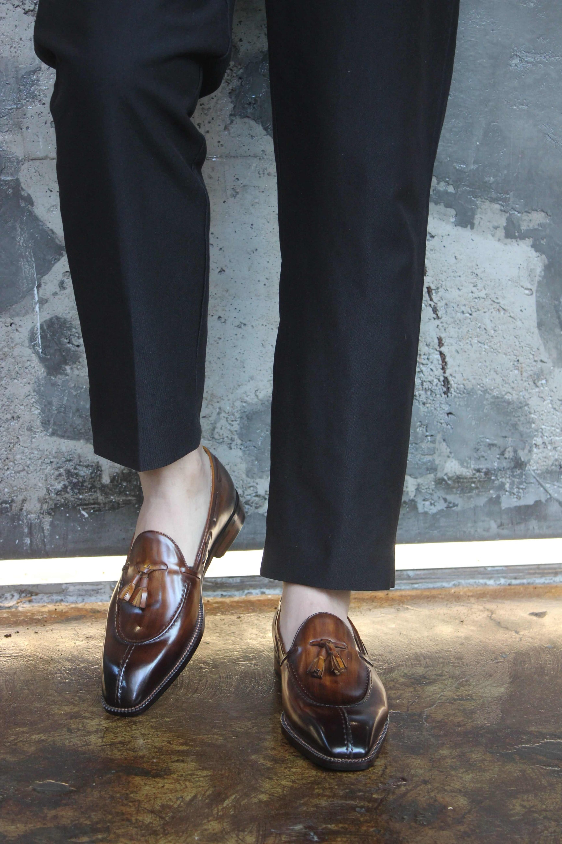 Hand-Burnished Brown Patina Leather Tassel Loafers – worn with cuffed black trousers, standing on a concrete floor, highlighting the elegant finish and tassel detail.