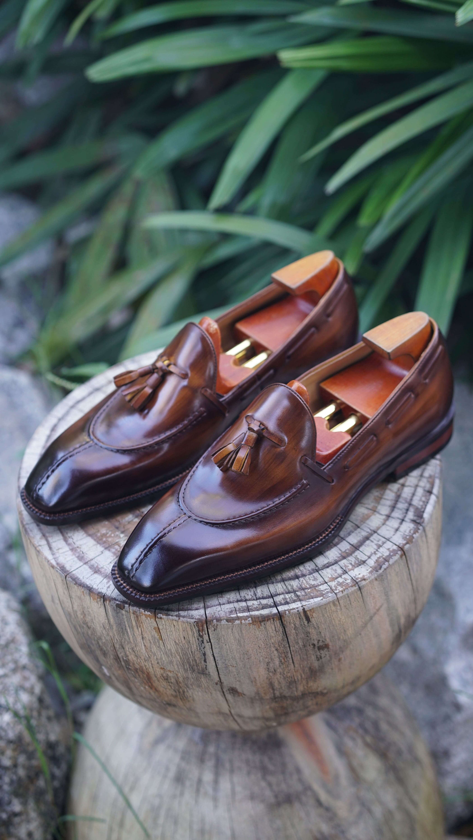 Hand-Burnished Brown Patina Leather Tassel Loafers – pair with shoe trees on a wooden stump, showcasing polished leather and classic tassel detail.