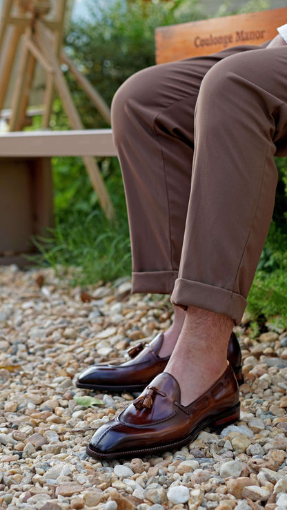 Hand-Burnished Brown Patina Leather Tassel Loafers – worn with cuffed brown trousers, sitting on a bench, highlighting rich patina and timeless style.