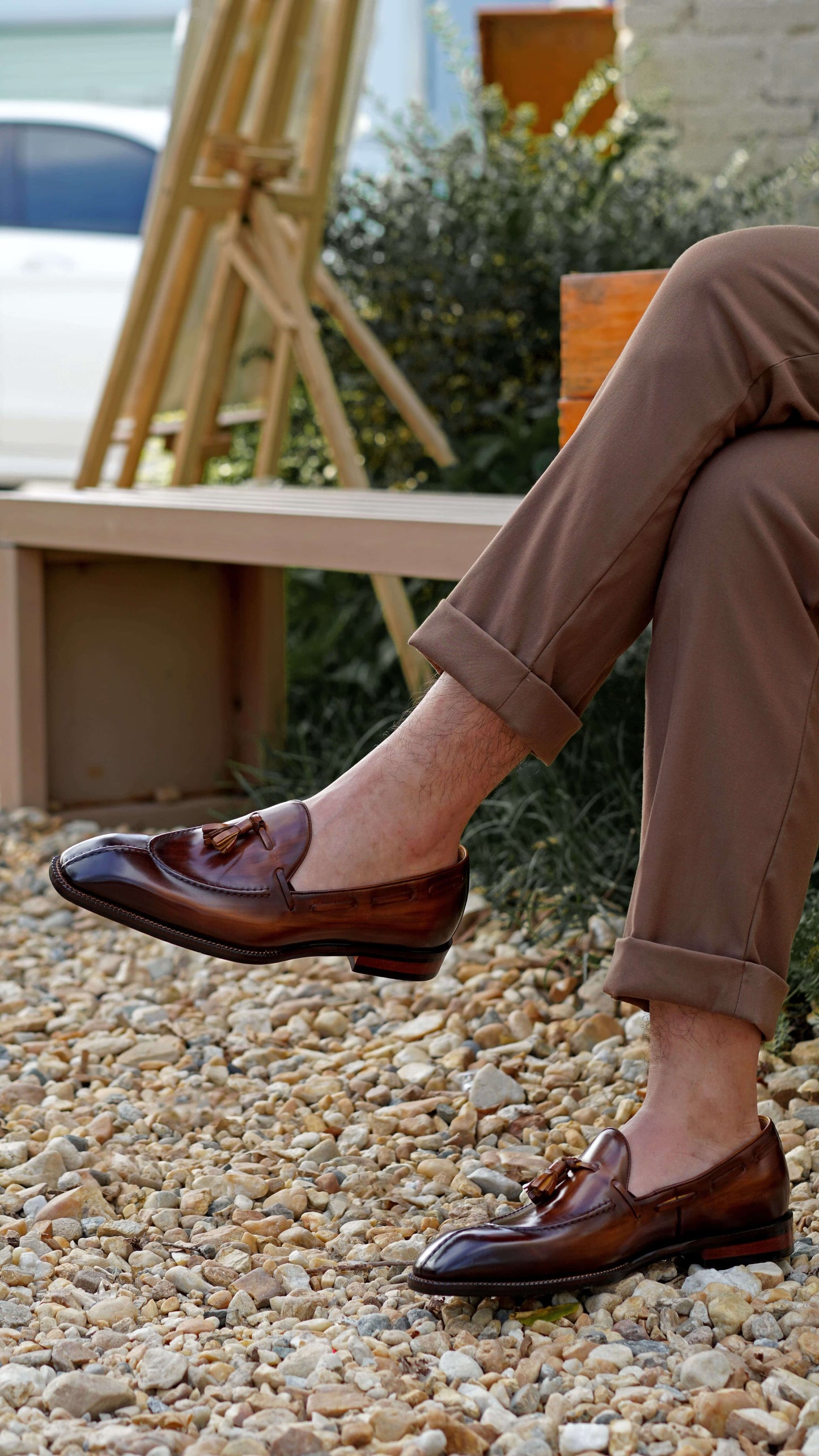 Hand-Burnished Brown Patina Leather Tassel Loafers – worn with cuffed brown trousers, sitting on a bench, showcasing polished leather and classic tassel detail.