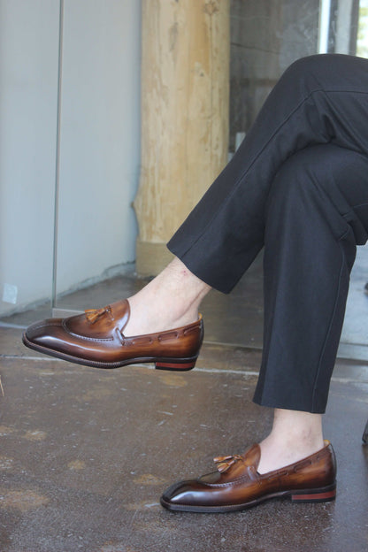 Hand-Burnished Brown Patina Leather Tassel Loafers – pair on a wooden stool, highlighting the hand-burnished patina and sleek almond-toe shape.