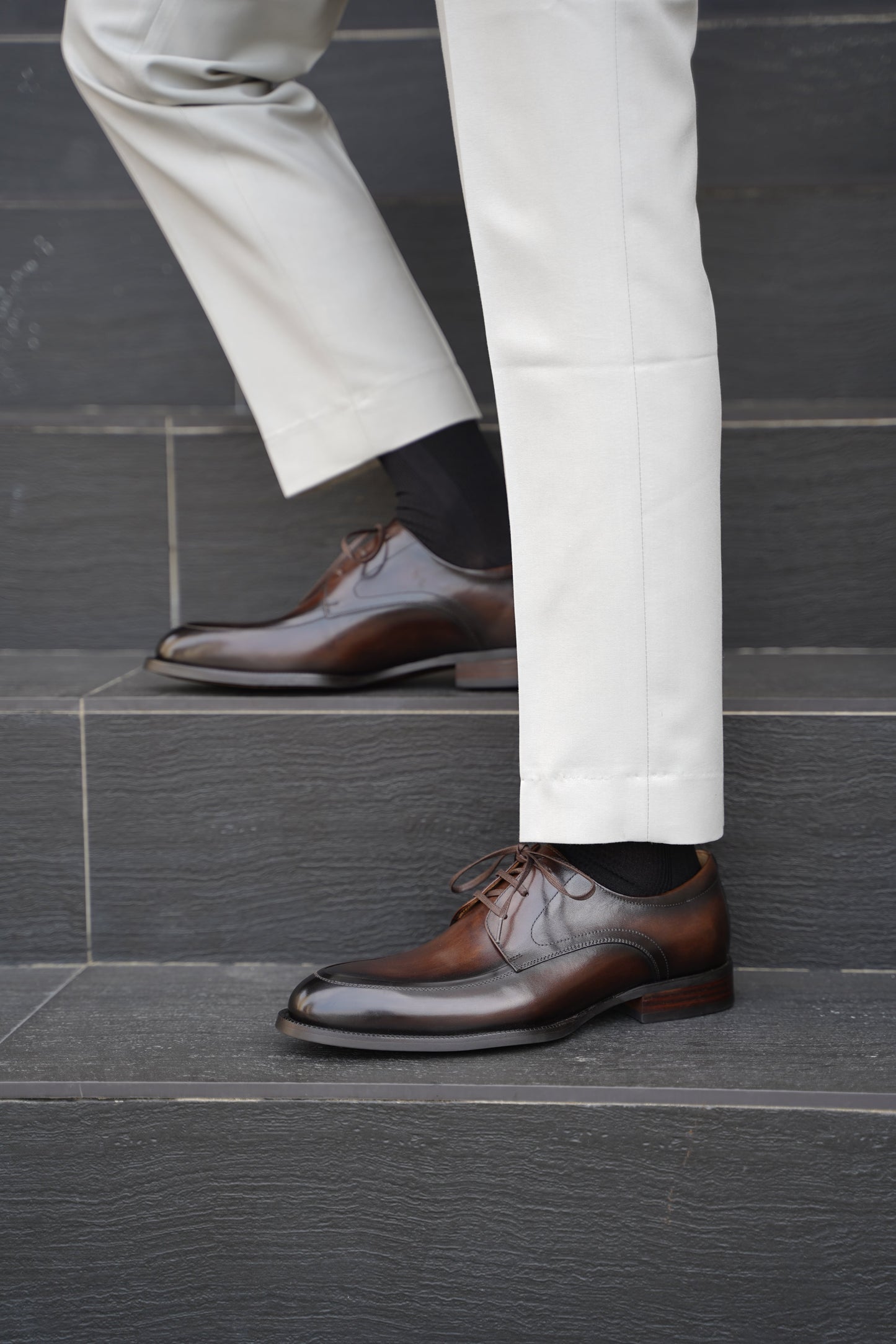 A man wearing Hand-Burnished Chestnut Split-Toe Derby shoes with light trousers, standing on steps outdoors.