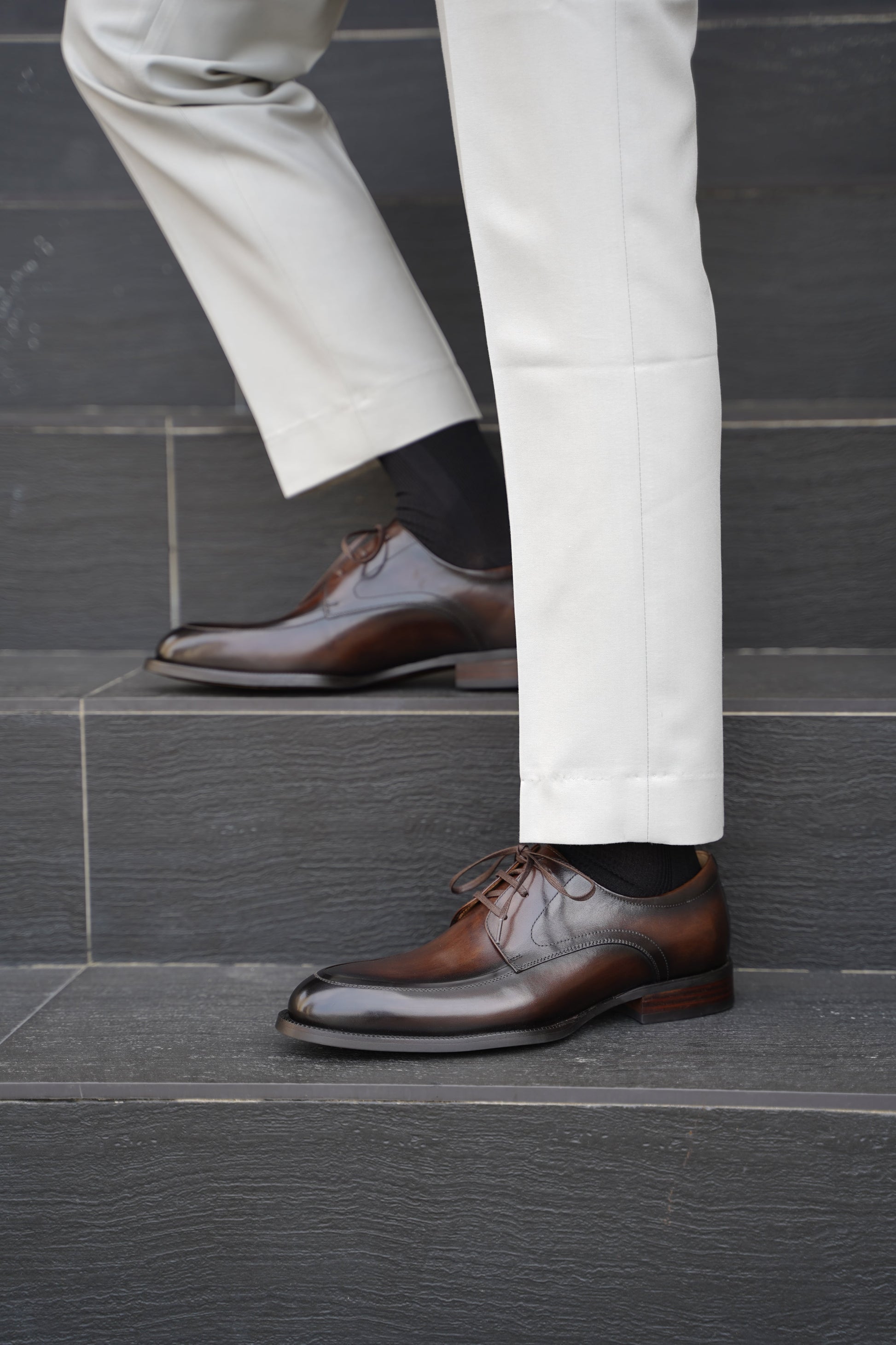 A man wearing Hand-Burnished Chestnut Split-Toe Derby shoes with light trousers, standing on steps outdoors.