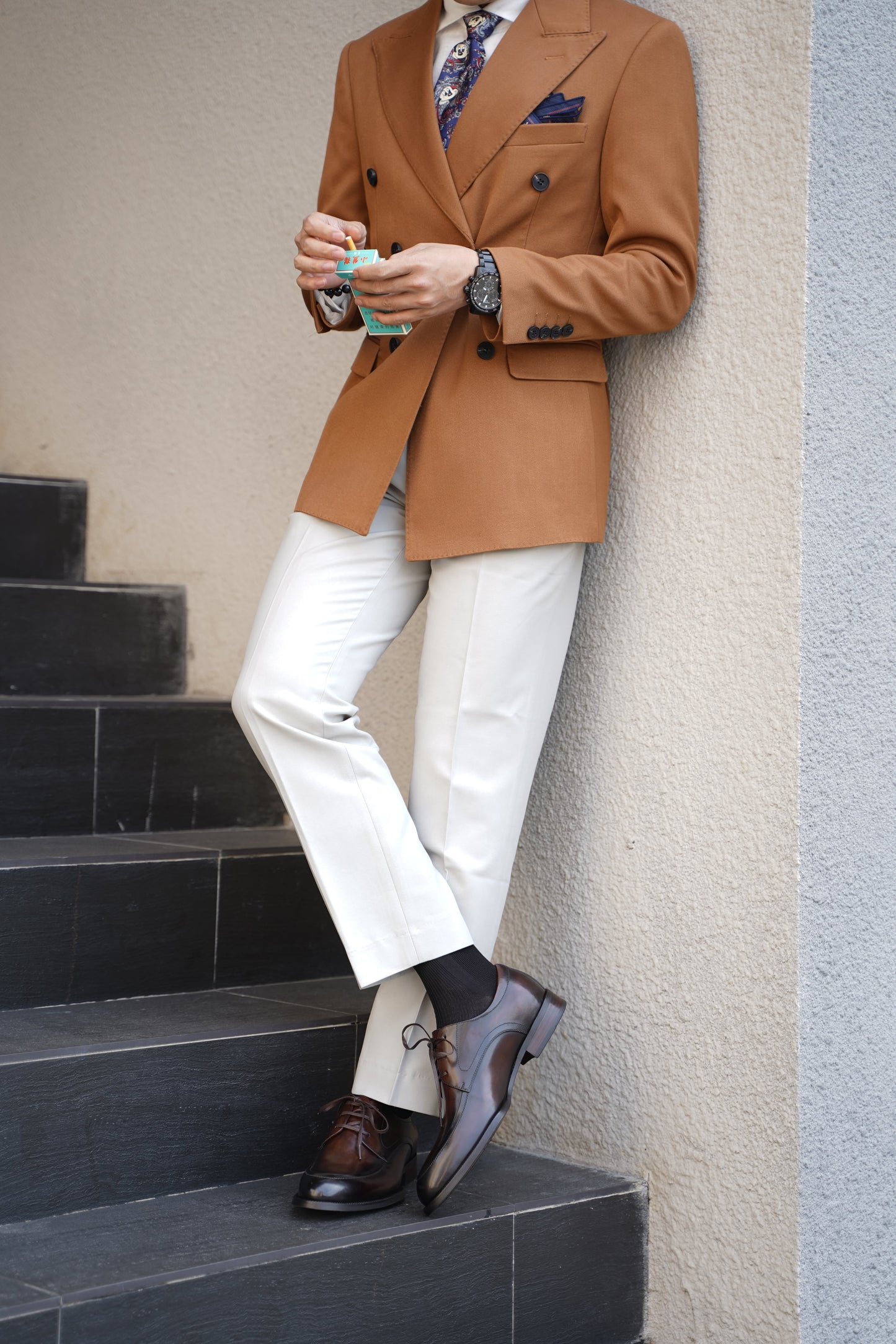 A man wearing HolloShoe Hand-Burnished Chestnut Split-Toe Derby shoes with light trousers and a brown blazer, leaning against a wall outdoors.