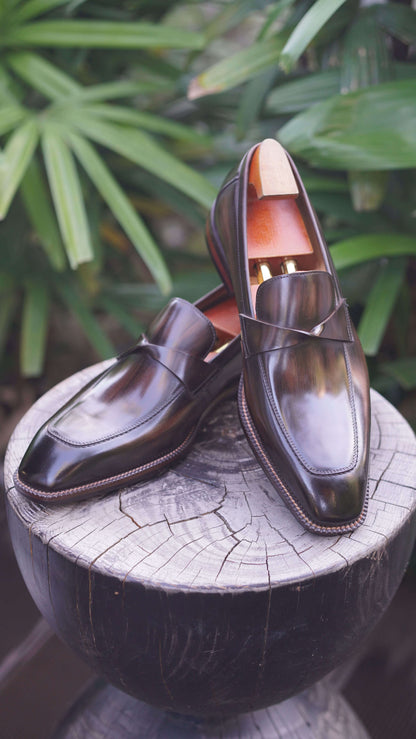 Hand-Painted Dark Brown Leather Penny Loafers – pair with shoe trees on a wooden stump, with green foliage, highlighting hand-painted patina and Italian craftsmanship.