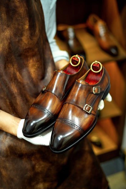 Hand-Polished Brown Double Monk Strap Shoes – held by gloved hands, showcasing rich marbled patina leather and polished double buckle closure.