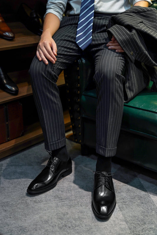 Man wearing Refined Black Derby Shoes – polished black leather, paired with pinstripe trousers and blue tie, sitting for work and leisure.