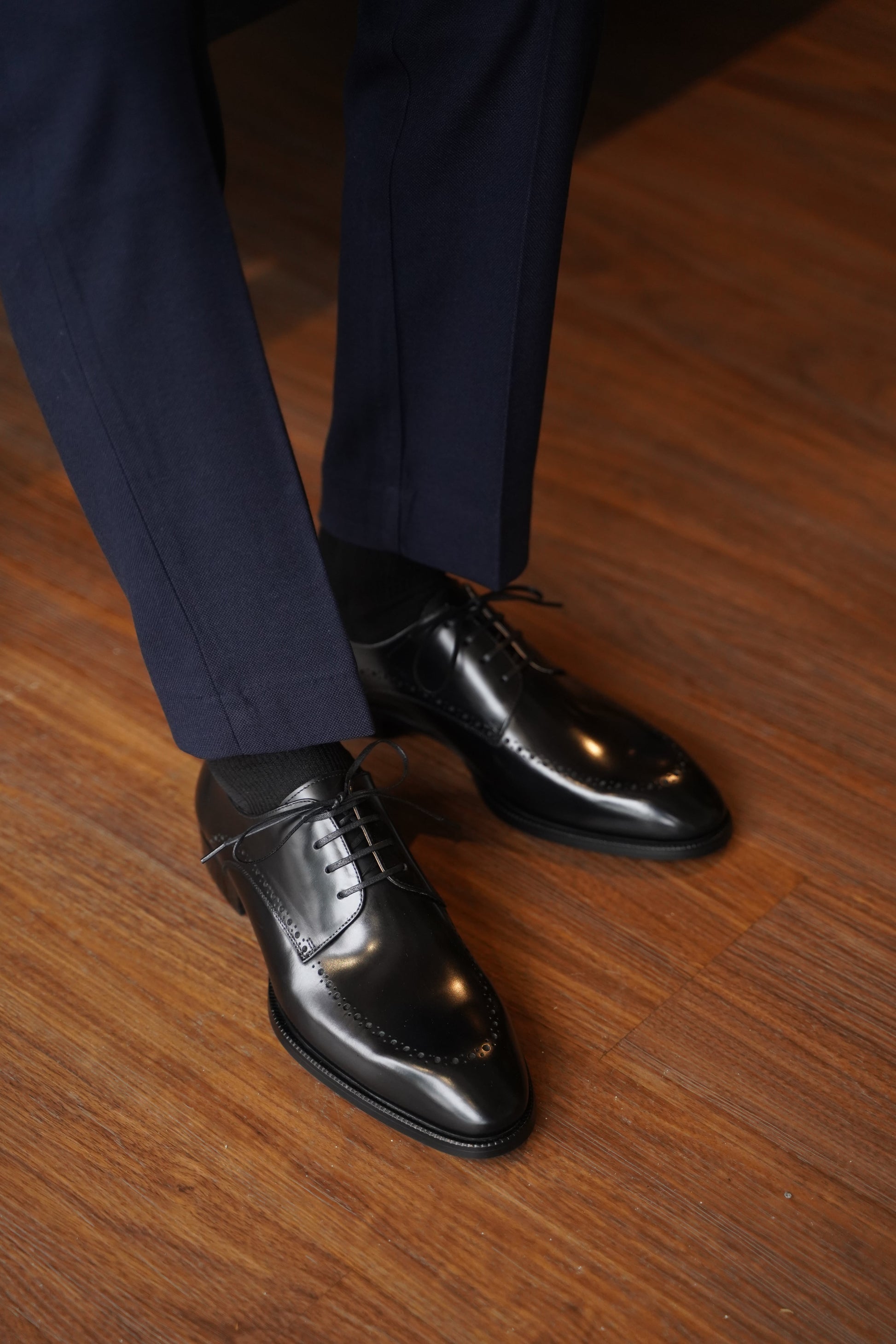 Man wearing Handcrafted Black Half-Brogue Derby Shoes – polished leather, paired with navy trousers on a wooden floor. 