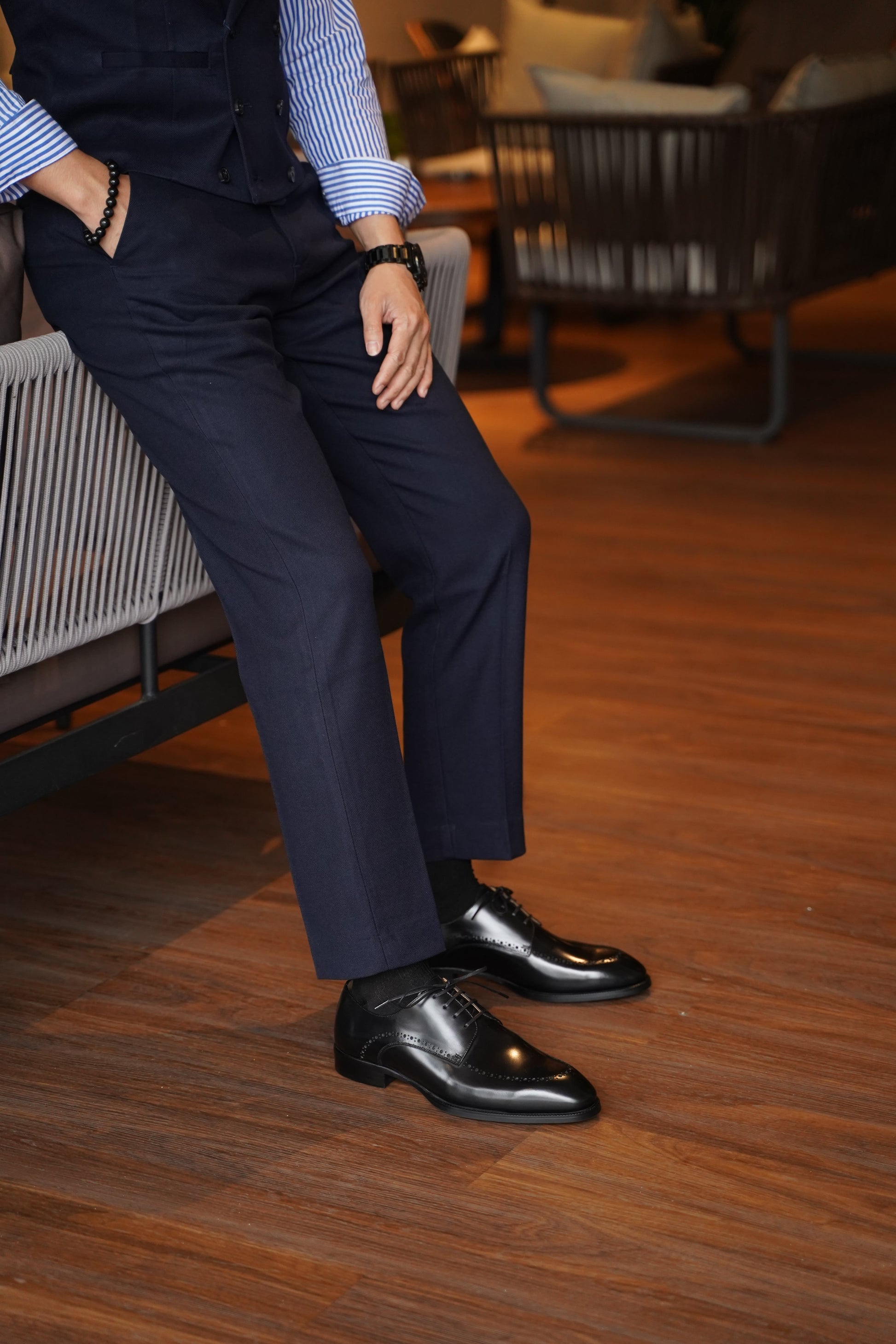 Man seated wearing Handcrafted Black Half-Brogue Derby Shoes – polished leather, paired with navy trousers for a smart formal look. 