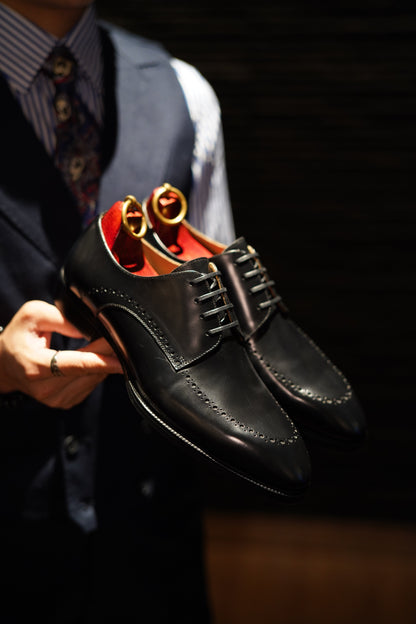 Handcrafted Black Half-Brogue Derby Shoes – high-shine polished leather, held by a man showcasing modern formality and red lining. 