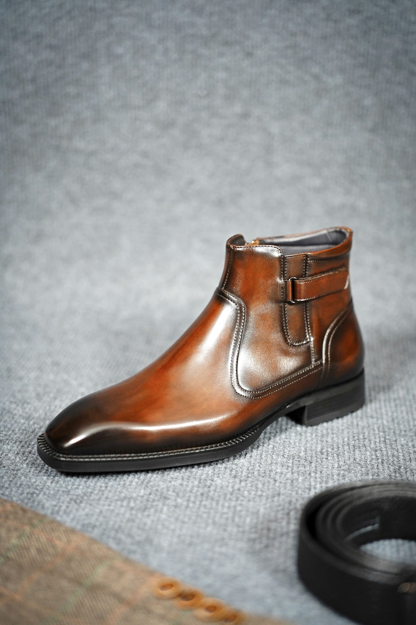 Handcrafted Brown Leather Zip-Up Ankle Boots with Buckle Detail – a close-up side view of a single boot on a gray textured background, highlighting the burnished finish.