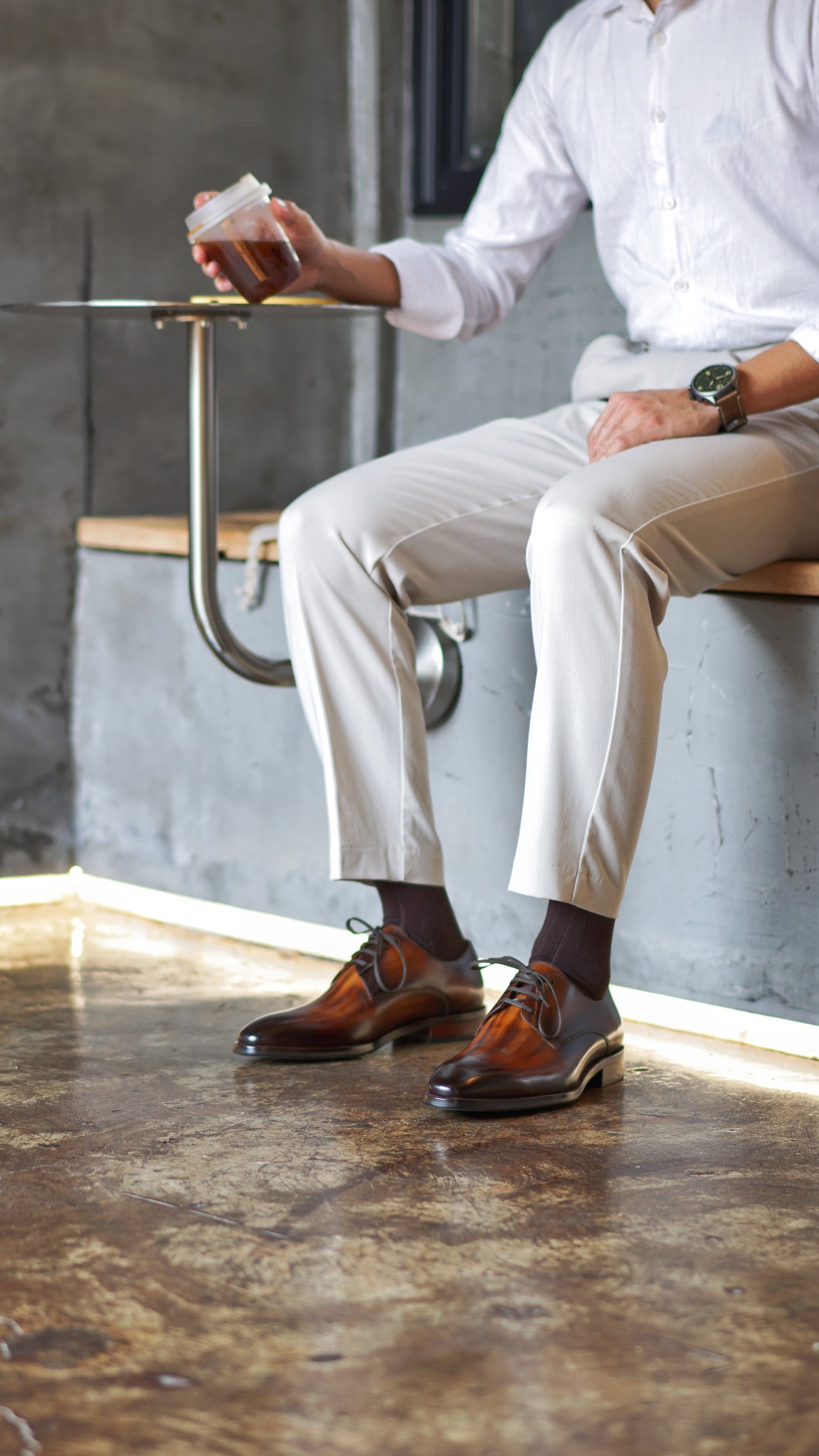 Man wearing hand-finished brown patina derby shoes and light grey cuffed trousers, sitting at a wooden table with a drink.