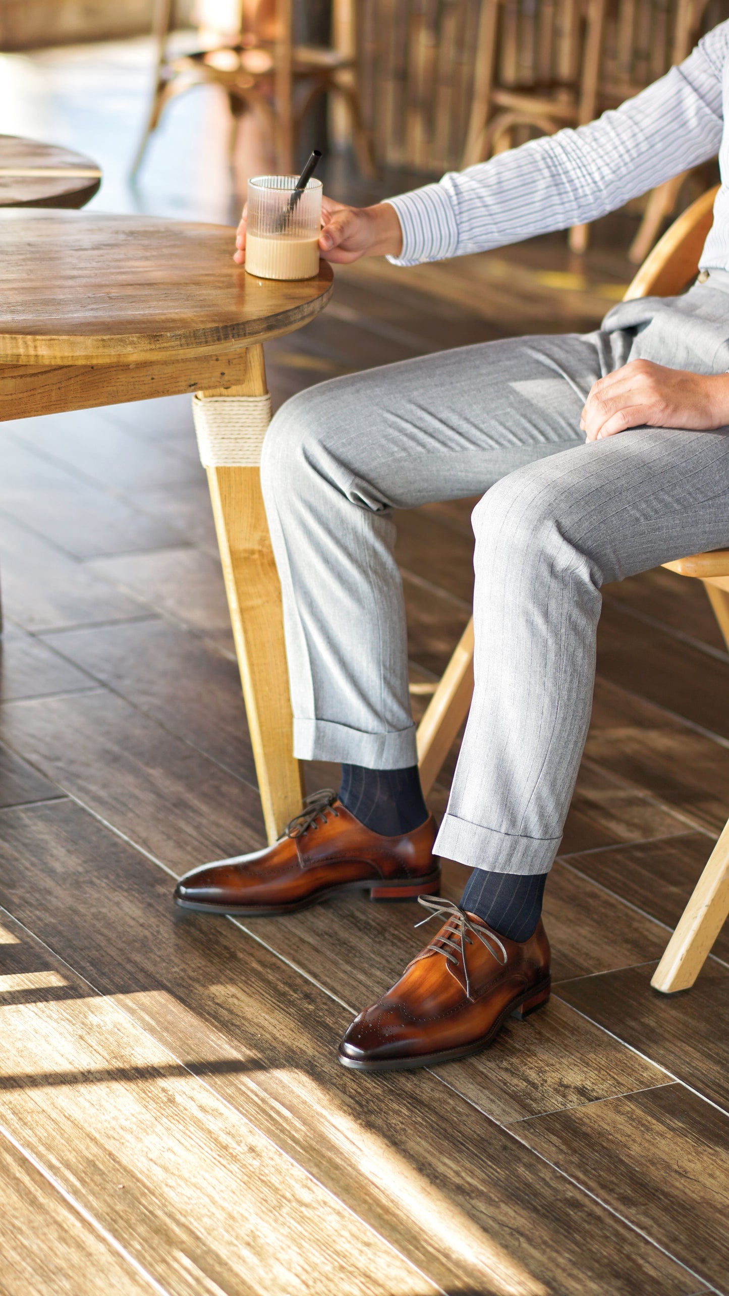 Man wearing brown patina derby shoes and beige trousers, sitting on a bench with a drink. 