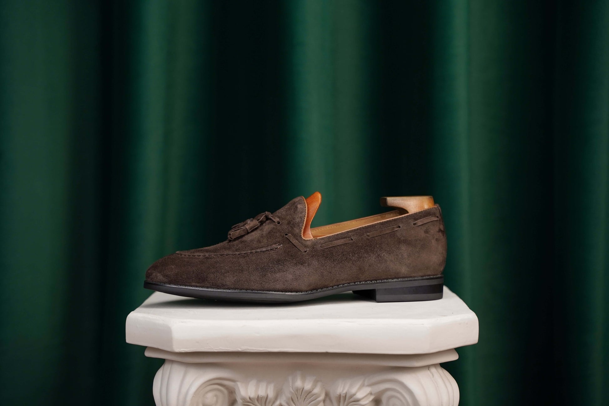 Handcrafted Umber Dark Brown Tassel Suede Loafer – side view with shoe tree, resting on a classical pedestal, highlighting artisanal finish and refined style.