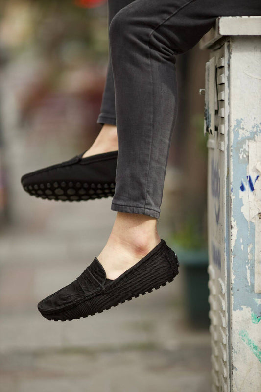 Handmade Black Knitted Driving Loafers – worn by a man in grey jeans, sitting on a utility box, showcasing the comfortable leather lining and casual style.