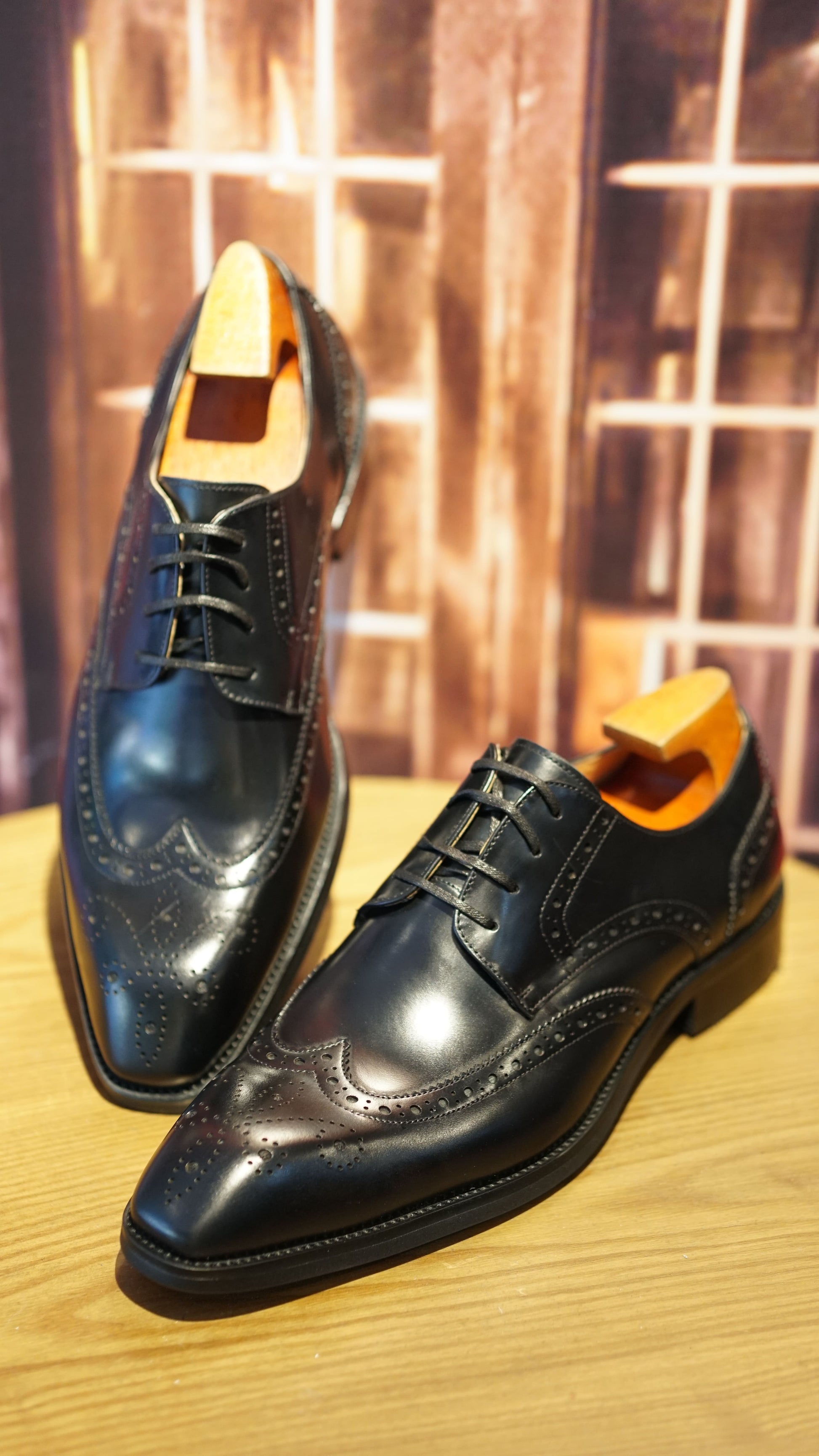 Handmade Black Wingtip Derby Shoes – elegant premium leather, pair displayed on a wooden surface at an angle, highlighting brogue details and shoe trees.