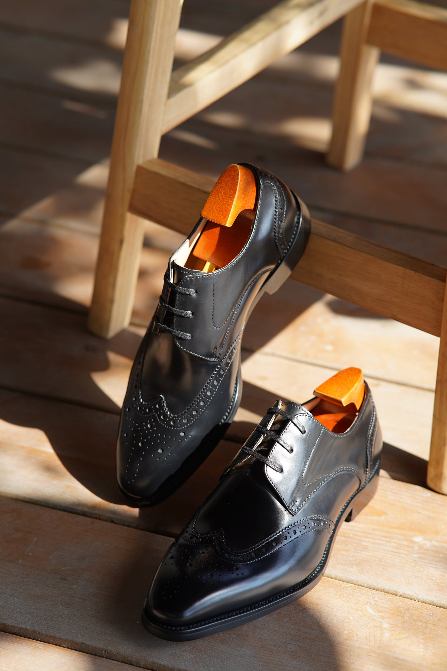 Handmade Black Wingtip Derby Shoes – premium polished leather, pair with shoe trees resting on a wooden chair in natural sunlight.