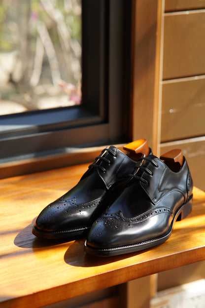 Handmade Black Wingtip Derby Shoes – elegant design in polished black leather, pair with shoe trees displayed on a wooden table by a window.