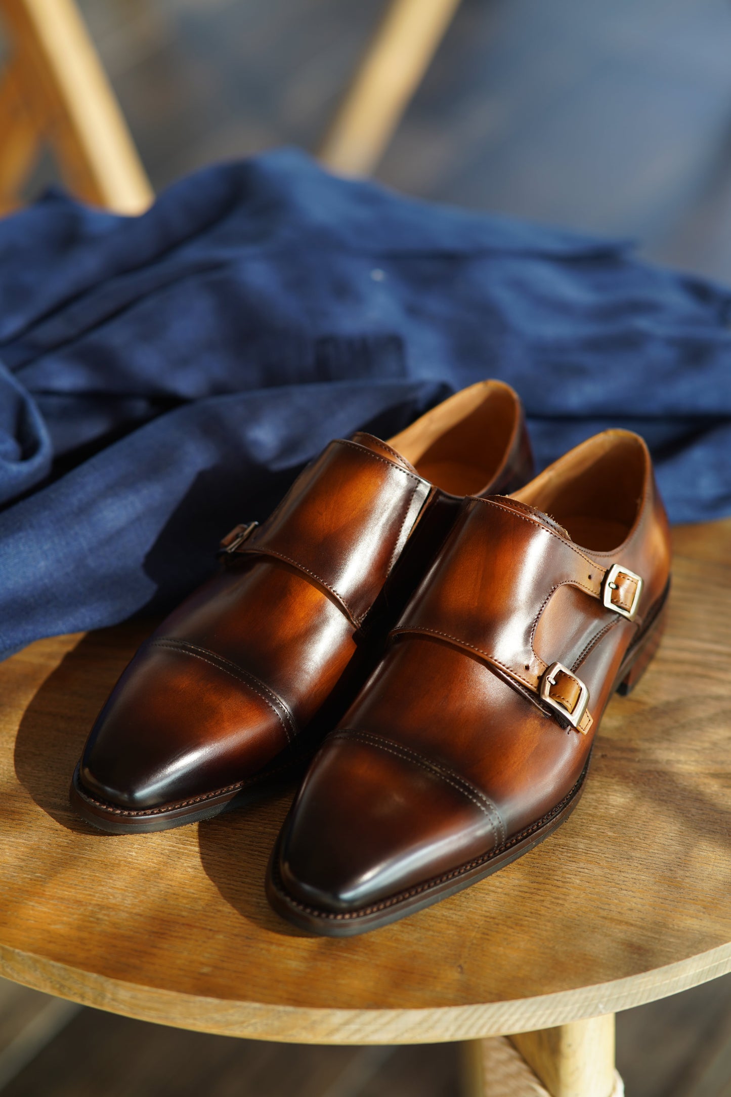 Handmade Brown Double Monk Strap Dress Shoes – pair on a wooden chair with blue fabric, highlighting rich patina finish and dual buckle closures.