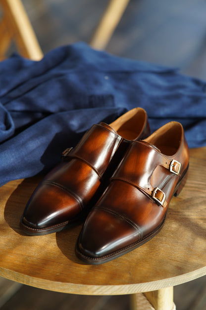 Handmade Brown Double Monk Strap Dress Shoes – pair on a wooden chair with blue fabric, highlighting rich patina finish and dual buckle closures.