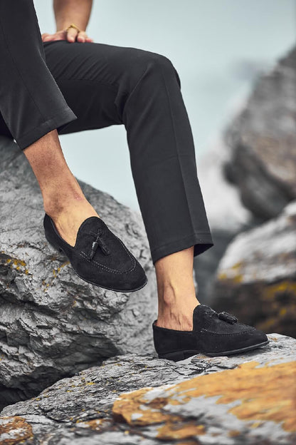 HolloShoe Black Suede Tassel Loafers - Sophisticated Leather Footwear – worn with black trousers, sitting on a rocky surface, showcasing timeless elegance and a refined aesthetic.