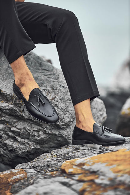 HolloShoe Black Tassel Loafers - Sophisticated Leather Footwear – worn by a man in black trousers, sitting on a rocky ledge, showcasing timeless design and elegance.