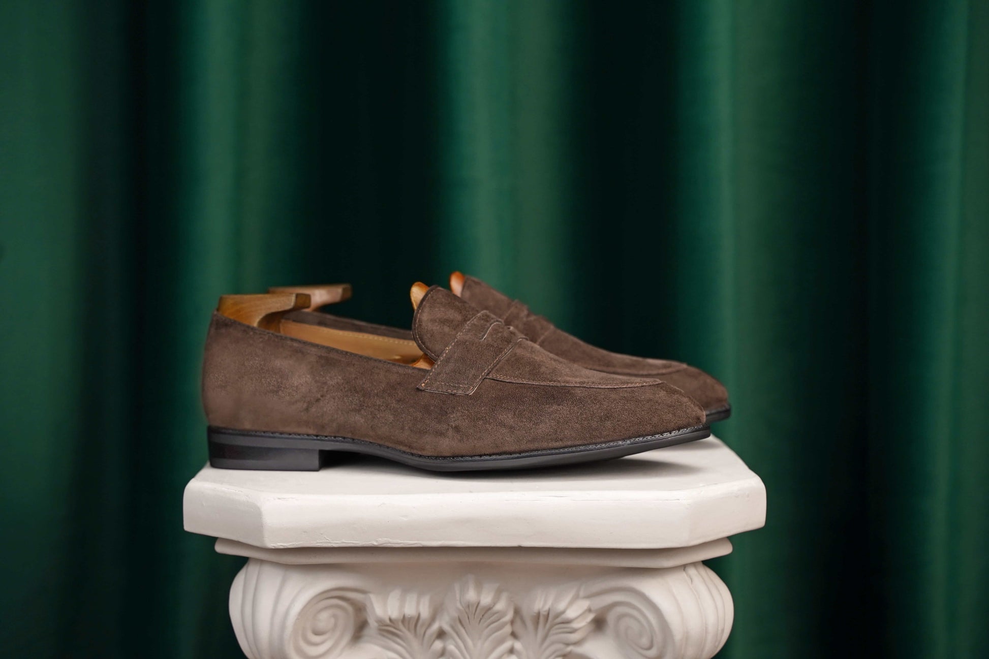 Earth Brown Suede Penny Loafers – pair with shoe trees, resting on a white classical pedestal, highlighting rich brown suede.