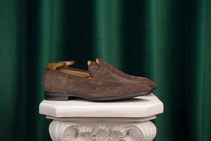 Earth Brown Suede Penny Loafers – pair with shoe trees, resting on a white classical pedestal, highlighting rich brown suede.