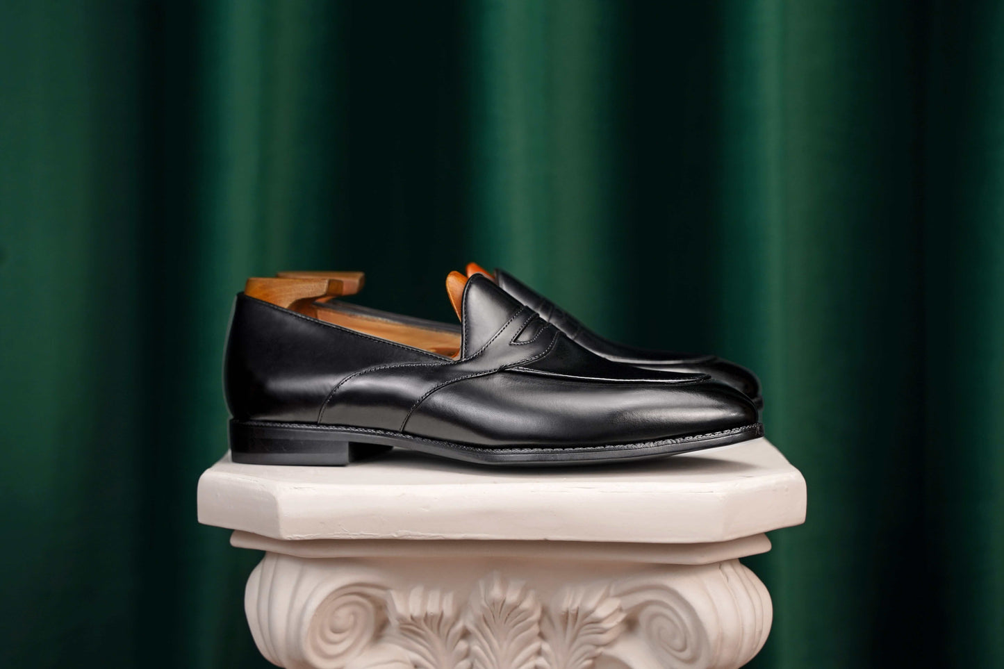 Handmade Black Leather Penny Loafers – pair with shoe trees, resting on a white classical pedestal against a green curtain, for elegant presentation.