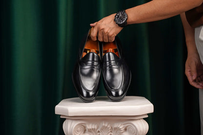 HolloShoe Handmade Black Leather Penny Loafers – pair held by a man's hand with a watch, showcasing sleek black leather and classic design against a green curtain.