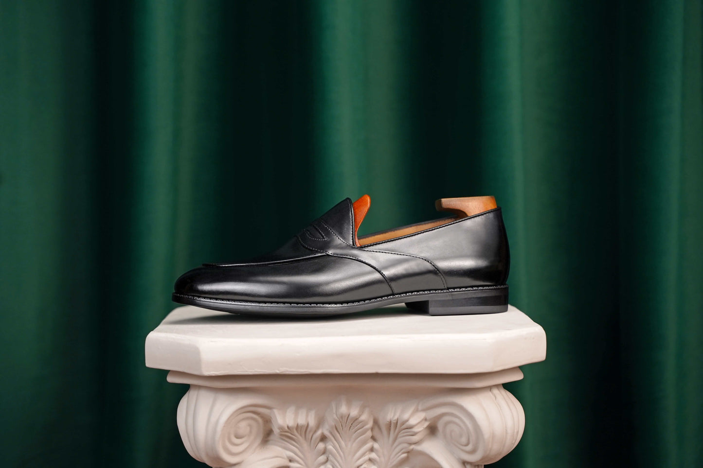 HolloShoe Handmade Black Leather Penny Loafer – side view with shoe tree, resting on a white classical pedestal against a green curtain, for elegant display.