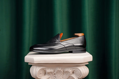 HolloShoe Handmade Black Leather Penny Loafer – side view with shoe tree, resting on a white classical pedestal against a green curtain, for elegant display.