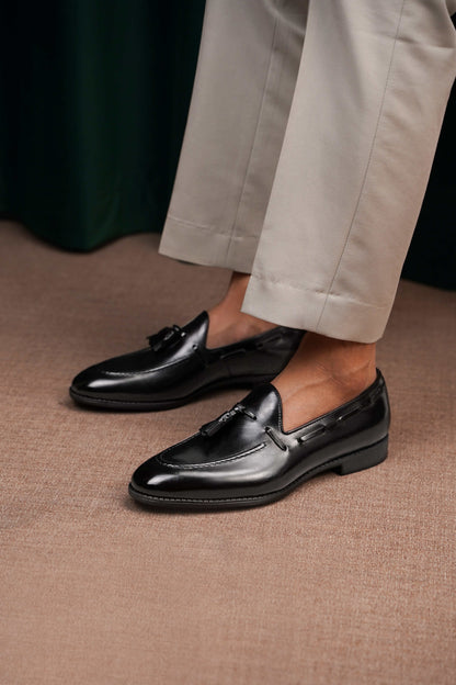 HolloShoe Handmade Black Tassel Leather Loafers – worn by a man in light trousers, sitting on a stone pedestal, highlighting effortless tailoring and quiet distinction.
